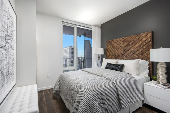 Modern bedroom with a large bed, white and gray bedding, and a wooden headboard. Two lamps on either side, large artwork on the wall, and a window showing a cityscape view.