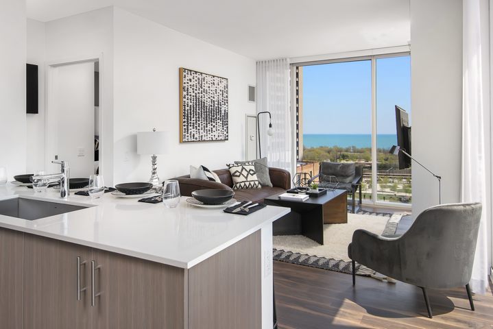Modern open-concept living room and kitchen with a large window offering a sea view. Features include a sectional sofa, wall art, kitchen island, and contemporary decor.