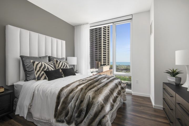 Modern bedroom with a large bed adorned with pillows and a faux fur throw. A floor-to-ceiling window reveals a cityscape and distant water view. A dresser and lamps are present.