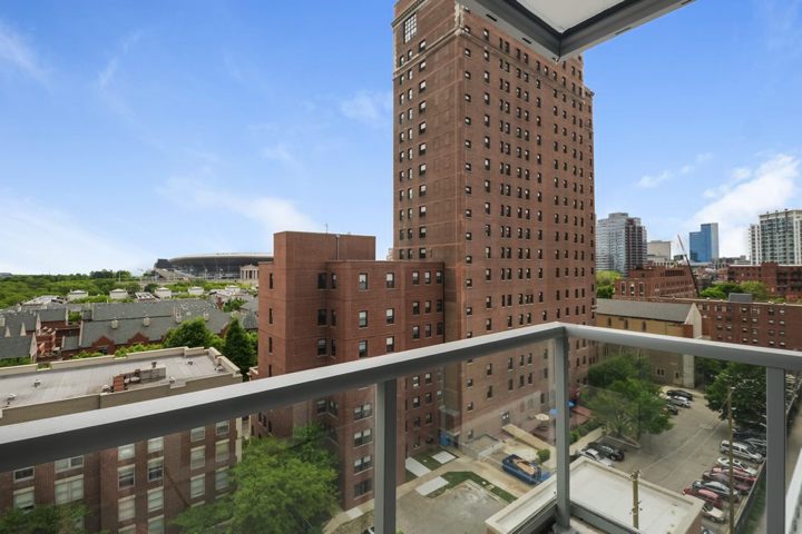 Cityscape view from a high-rise balcony showing brick buildings, greenery, and modern skyscrapers under a clear blue sky.
