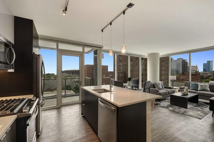 Modern open-concept kitchen and living area with dark cabinets, island, and large windows showing a cityscape view. Two pendant lights hang above the island.