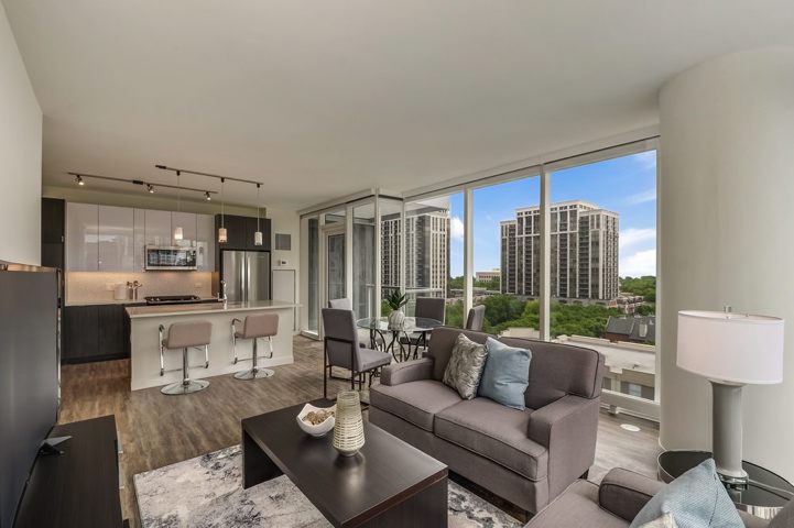 Modern living room with gray furniture, open kitchen with bar stools, and large windows showing cityscape and trees.