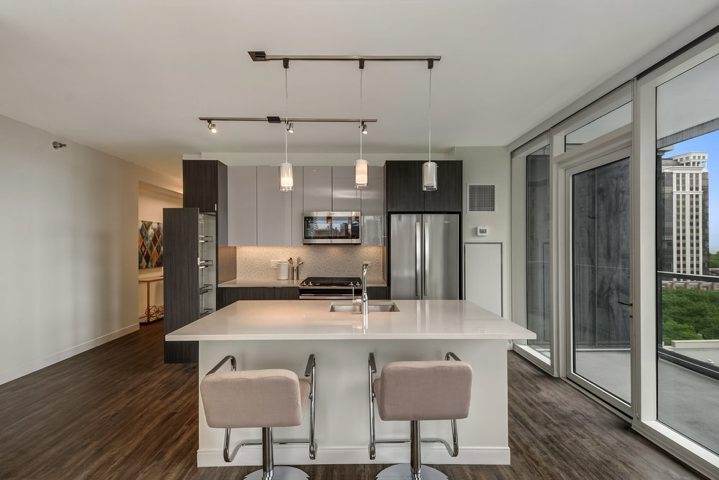 Modern kitchen with white island, two beige stools, dark wood cabinets, stainless steel appliances, and large windows offering a city view.