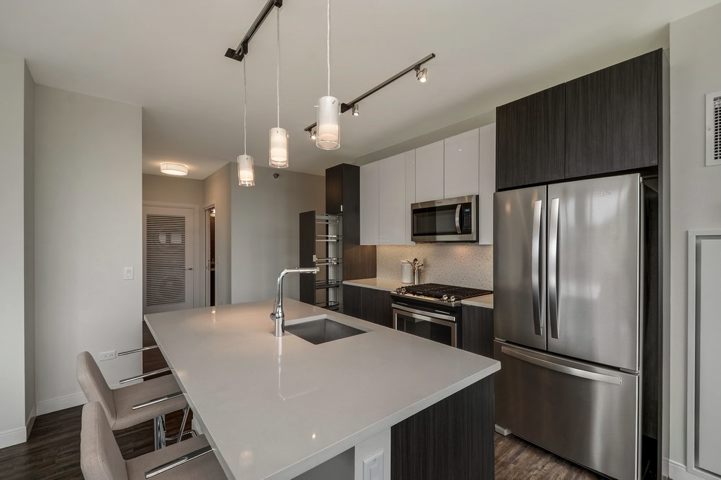 Modern kitchen with dark cabinets, stainless steel appliances, and a gray quartz island with a sink. Pendant lights hang above the island. Two beige bar stools are positioned at the island.