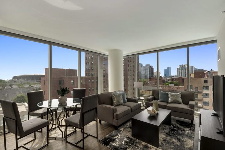 Modern living room with large windows showing a cityscape. Furnished with gray sofas, a black coffee table, round dining table, and chairs. Decor includes cushions and a potted plant.