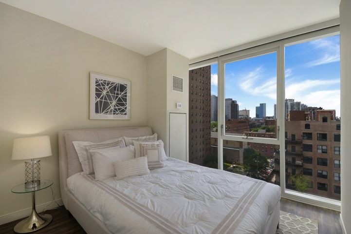 Bedroom with a neatly made bed, large windows showcasing a cityscape, a lamp on a side table, and wall art above the bed.