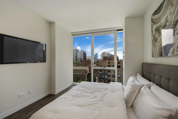 Modern bedroom with a bed, wall-mounted TV, abstract artwork, and large window overlooking city buildings.