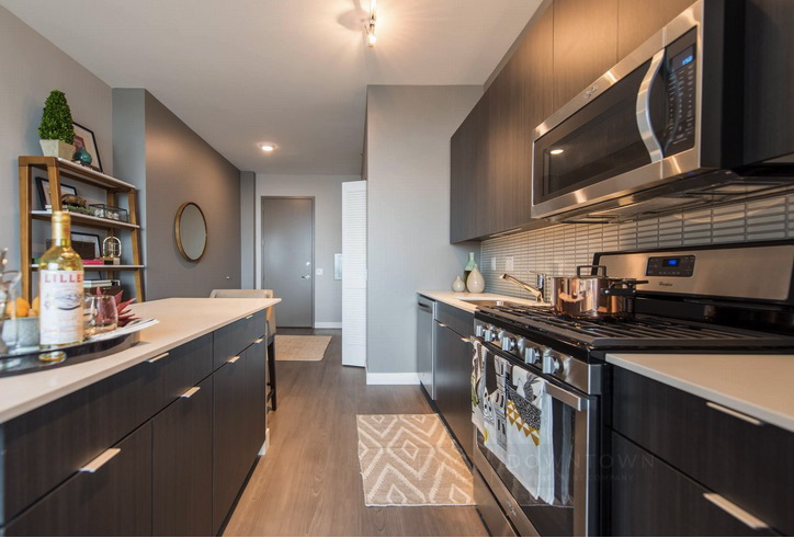 Modern kitchen with dark cabinets, stainless steel appliances, and a light wood floor. A rug is in front of the stove, and shelves with decor are on the left. A round mirror hangs on the wall.