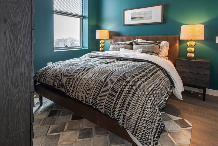 Bedroom with teal walls, a large bed with patterned black and white bedding, two bedside tables with golden lamps, and a window with a city view.