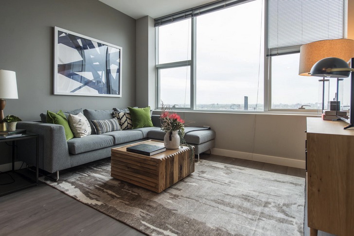 Modern living room with a grey sectional sofa, green and white pillows, wooden coffee table, abstract wall art, and large windows with blinds. A lamp and potted plant are also present.