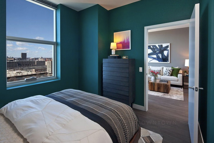 A bedroom with teal walls, a bed, a tall dresser, and a window with a city view. An open door leads to a living room with a couch and artwork on the wall.