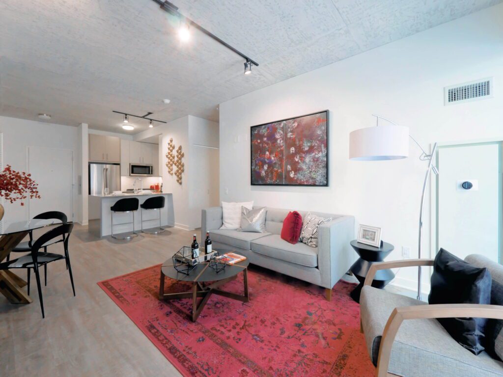 Modern living room with a gray sofa, red rug, and a coffee table. Kitchen with bar stools visible in the background. Contemporary artwork and floor lamp add decorative elements.