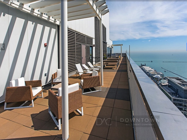 Rooftop terrace with wicker furniture, white cushions, and sun loungers; overlooking a body of water and city skyline.