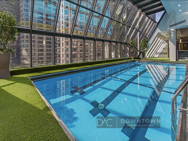 Indoor swimming pool with clear water, surrounded by artificial grass, located in a glass-enclosed area with a view of city buildings.
