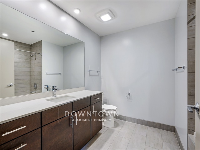 Modern bathroom with wooden vanity, white countertop, and a toilet. Wall-mounted towel bars on both sides, large mirror, and recessed lighting above. Grey tile flooring.