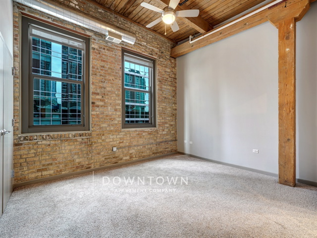 Empty room with exposed brick walls, wooden beams, carpeted floor, and two large windows. Ceiling fan and industrial ductwork are visible.