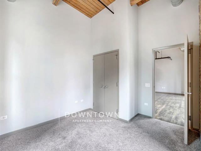 Room with carpet flooring, light gray walls, wooden ceiling beams, and an open door leading to another room. Two closed gray doors are on the left wall.