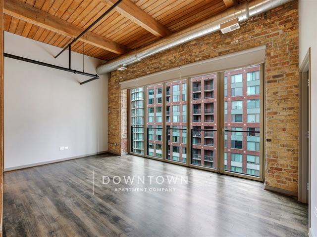 Loft-style room with exposed brick wall, wooden ceiling, ductwork, and large windows overlooking a building. Dark wood flooring and a metal ceiling fixture are present.