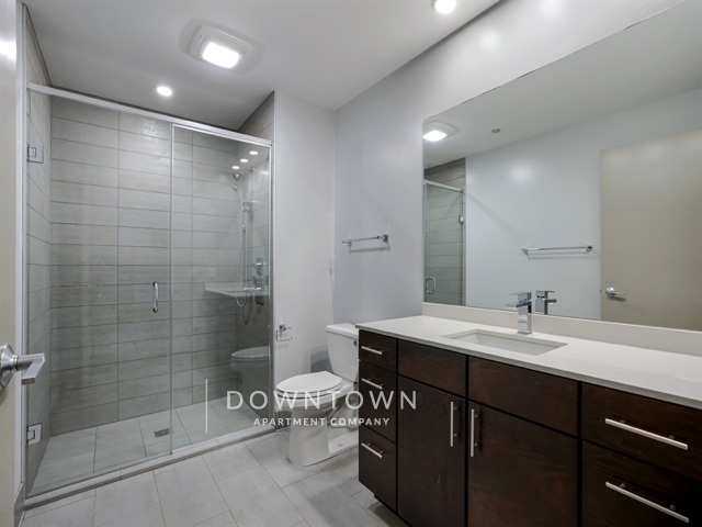 Modern bathroom with a glass-enclosed shower, toilet, and dark wood vanity with a sink. Bright lighting and neutral-colored tiles.