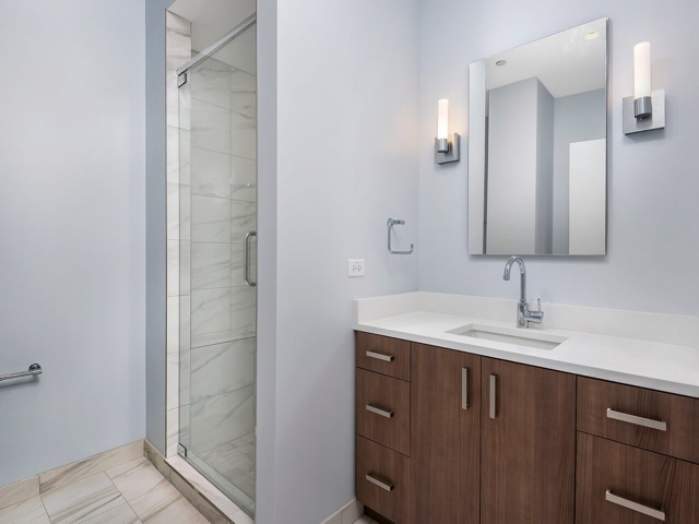 Modern bathroom with light wood vanity, white countertop, rectangular mirror, and wall-mounted lights. Glass-enclosed shower with tiled walls is visible on the left.