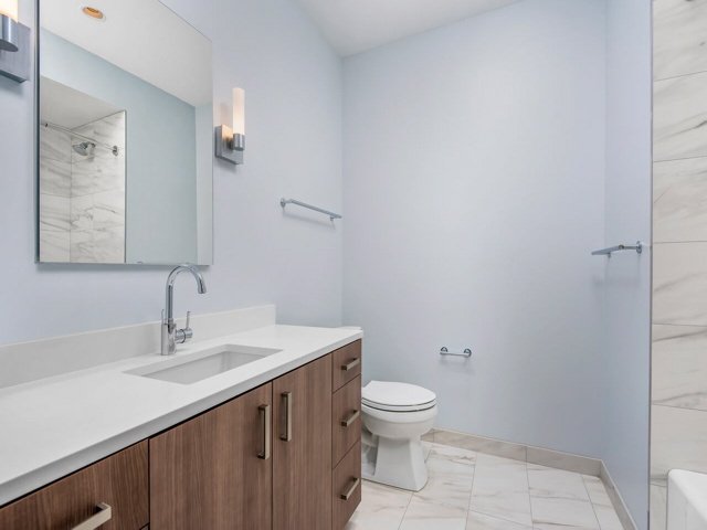 Modern bathroom with a wooden vanity, white countertop, wall-mounted mirror, toilet, and light gray walls. Tile flooring and wall-mounted lights are also visible.