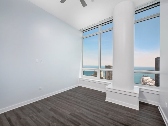 Empty room with light gray walls, large windows offering a city and water view, and dark wood flooring. A white column is near the corner.