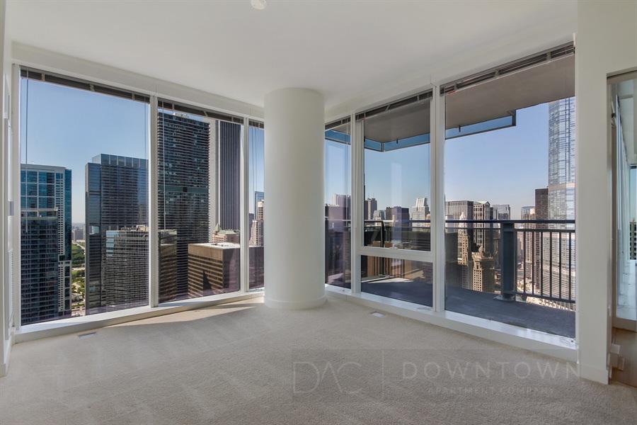Modern high-rise apartment interior with floor-to-ceiling windows and a city view. Beige carpet covers the floor, and a large balcony is visible through the glass doors.