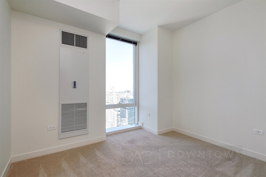 Empty room with beige carpet, large window showing a cityscape view, and a wall vent panel. Walls are white, and the space is well-lit.
