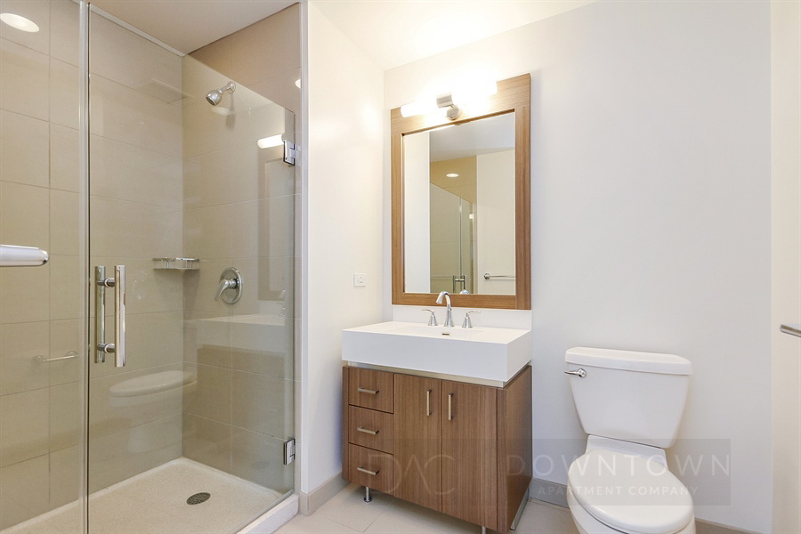 Modern bathroom with glass-enclosed shower, wooden vanity with sink and mirror, and a toilet. White walls and beige tile flooring create a clean, minimalist look.
