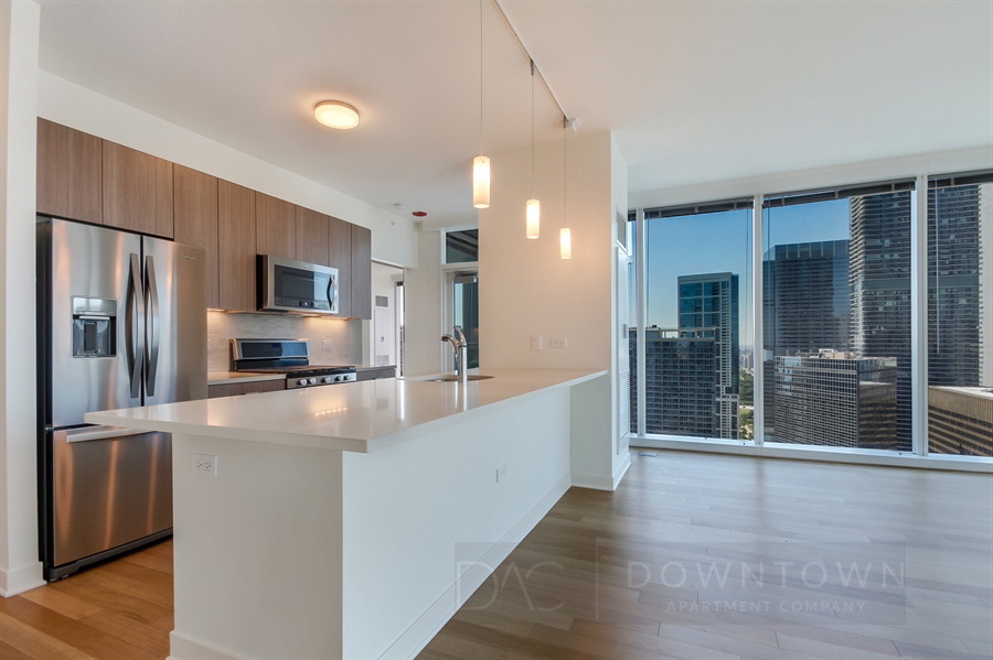 Modern kitchen with stainless steel appliances, white countertops, and wood cabinetry. Floor-to-ceiling windows show a cityscape view. Pendant lights hang above the island.