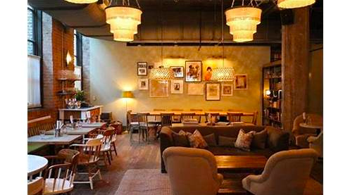 Cozy café interior with wooden tables, chairs, sofas, and warm lighting. Framed pictures adorn the walls, and beige lamps hang from the ceiling.