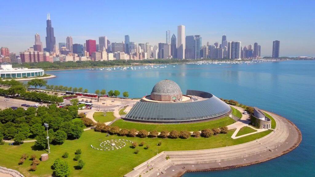Aerial view of a lakeside building with a dome, surrounded by green lawns and trees, located near a body of water. City skyline with tall buildings in the background under a clear blue sky.