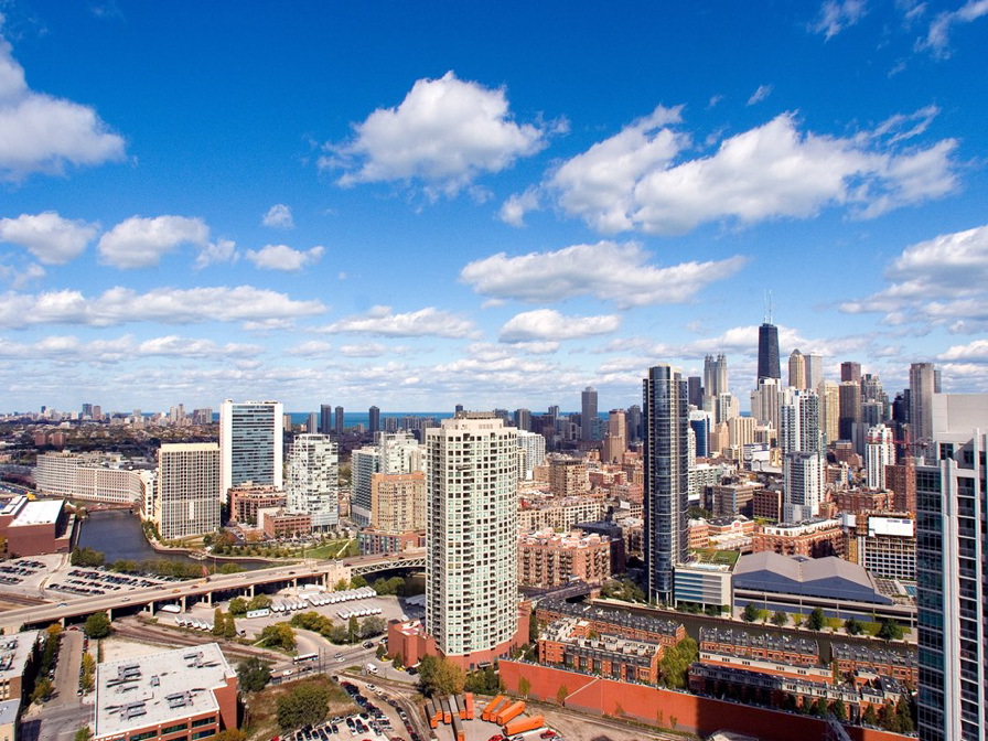 Aerial view of a cityscape with tall buildings, a river, and scattered clouds in a blue sky.