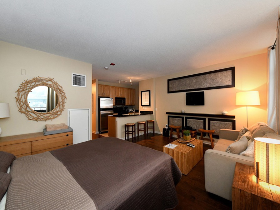 Studio apartment with bed, seating area, and kitchen. Features a round mirror, wall-mounted TV, sofa, and bar stools. Decor includes wood accents and soft lighting.