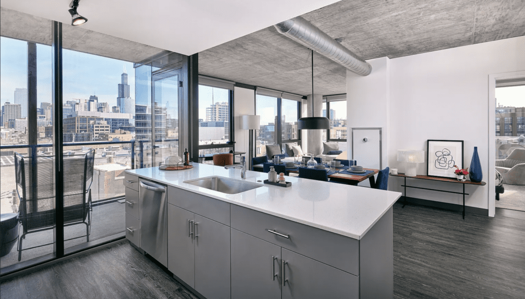 Modern kitchen and living area with cityscape view. Features include a kitchen island with sink, dining table, artwork, and floor-to-ceiling windows. Urban skyline visible outside.