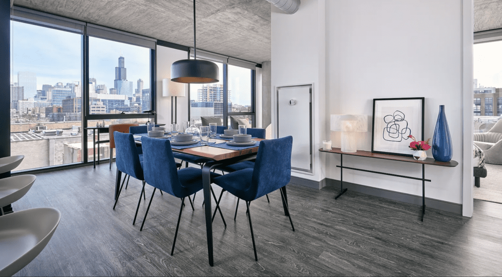 Modern dining room with blue velvet chairs, set table, and large windows offering a city skyline view. A minimalist side table displays art and decor under a concrete ceiling.