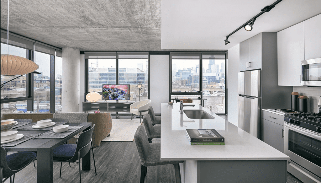 Modern open-plan kitchen and living area with city views. Features a kitchen island, dining table, sofa, TV, large windows, and visible urban skyline. Light and airy design.