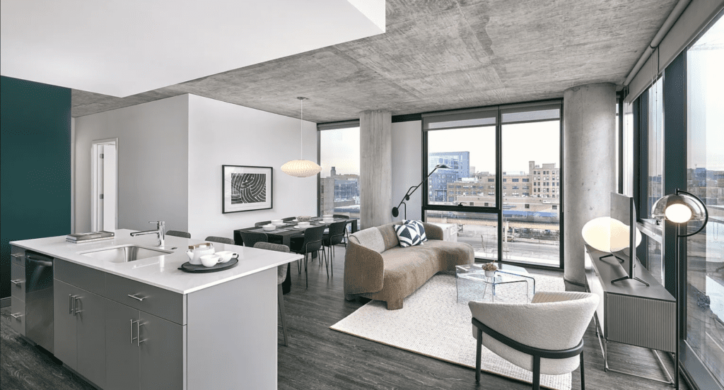 Modern apartment interior with an open-plan layout. Features include a kitchen island, dining table, and cozy living area. Large windows offer a view of city buildings in the background.