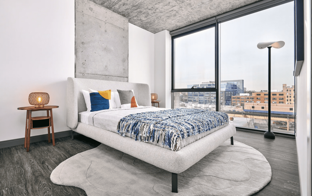 Modern bedroom with a large bed, decorative pillows, and a blue throw blanket. The room features a concrete accent wall, large windows with a city view, and a standing lamp next to the bed.