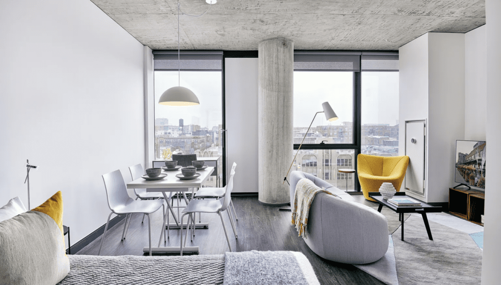 Modern apartment with open-plan layout featuring a living area, dining table, and bed. Large windows offer a city view. Decor includes a yellow chair, floor lamp, and concrete details.