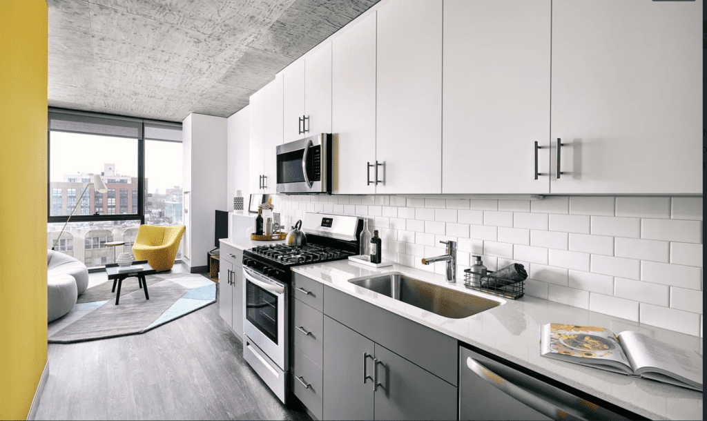 Modern kitchen with white cabinets, a stainless steel oven, and a microwave. Yellow accent wall, a grey couch, a yellow chair, and large windows are visible in the adjacent living area.