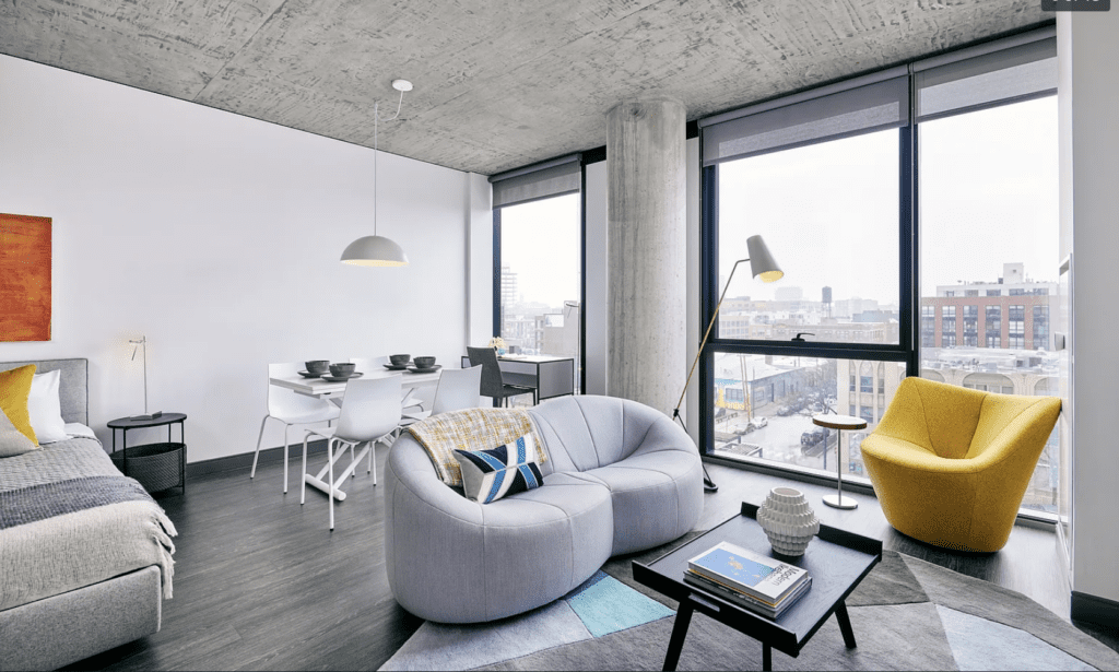 Modern apartment with concrete ceiling, featuring a bed, small dining table, gray sofa, yellow chair, and large windows overlooking a cityscape.