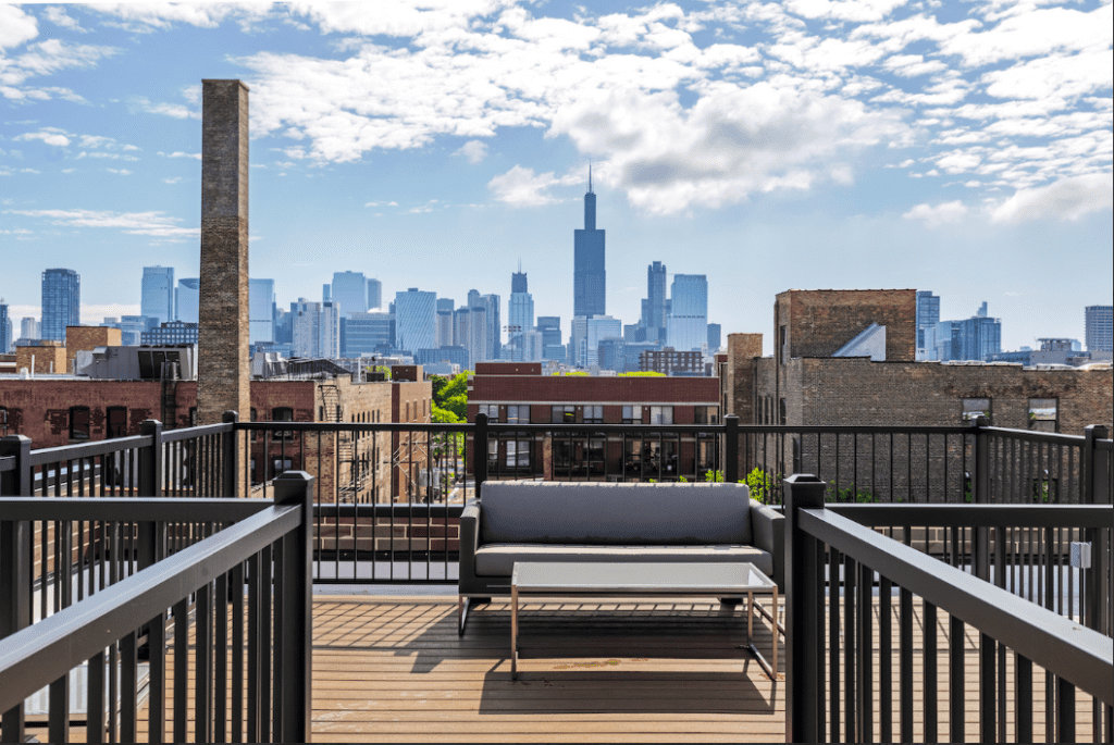Rooftop patio with a couch and table, overlooking a city skyline with a tall building in the distance under a partly cloudy sky.