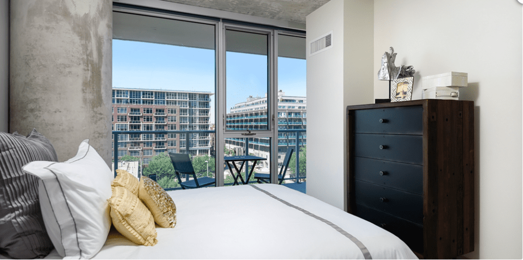 A bedroom with a neatly made bed, pillows, a wooden dresser, and a balcony view showing buildings outside on a clear day.