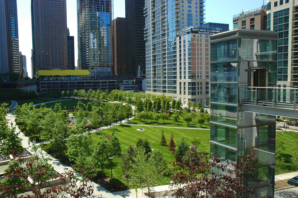 Urban park with green lawns and trees surrounded by tall buildings under a clear blue sky. There's a glass elevator structure on the right side of the image.
