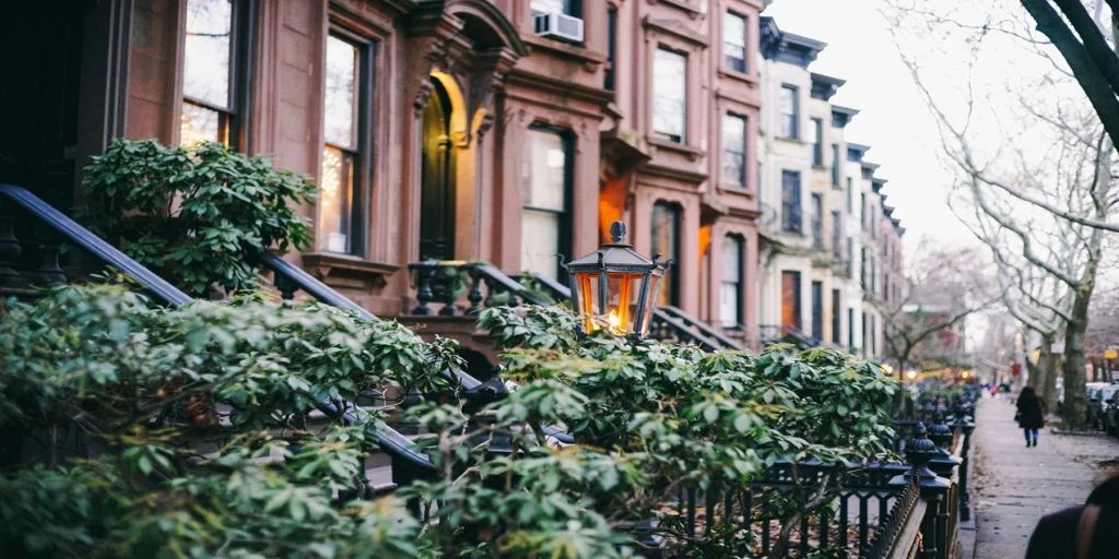 Street view of brownstone buildings with elegant staircases and manicured green shrubs, featuring a vintage streetlamp. Trees line the sidewalk in this slice of Chicago luxury real estate, where people stroll by in the distance.