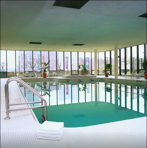 Indoor swimming pool with clear water, stainless steel handrail, and folded white towels on the tile floor. Glass walls reveal an outside view of buildings and plants surrounding the pool.