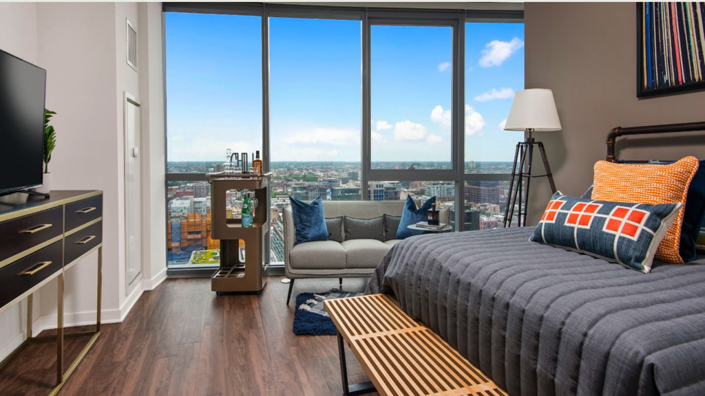 Modern bedroom with a large window offering a city view. Features a bed, bench, dresser, TV, and seating area. Decor is contemporary with warm tones and geometric cushions.