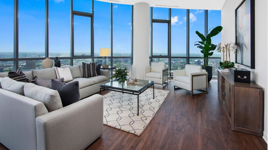 Modern living room with large windows, light gray sofas, a glass coffee table, and a geometric rug. Wooden floor, city view, tall plants, and a sideboard with framed art on the wall.