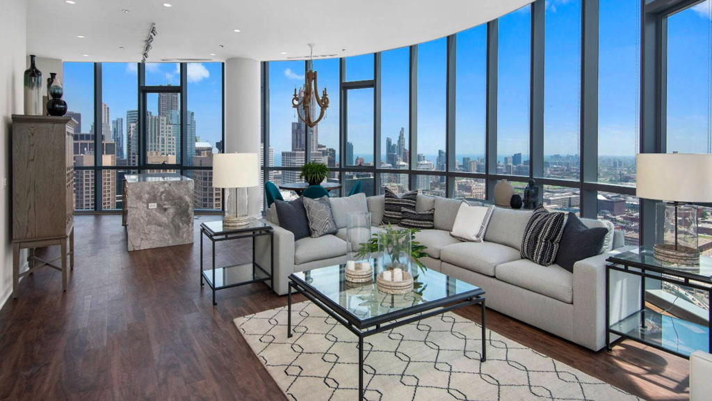 Modern living room with floor-to-ceiling windows showcasing a cityscape. Features a gray sectional sofa, glass coffee table, and decorative plants on a wooden floor.